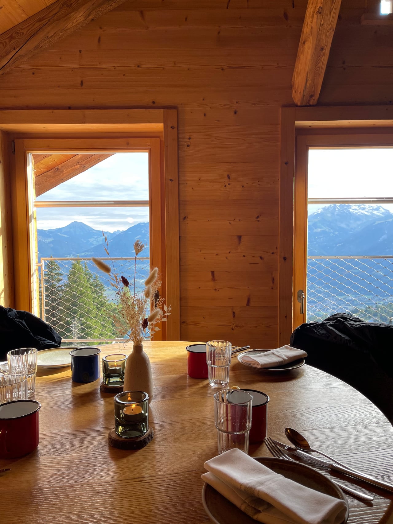 A chalet breakfast table set with glasses and dried flowers beside windows framing the mountains